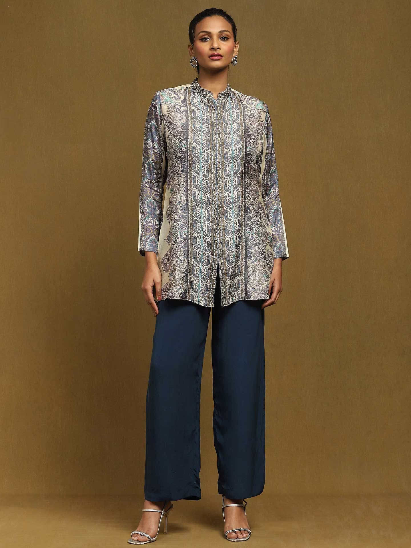 Woman wearing a patterned top and navy pants on a brown background