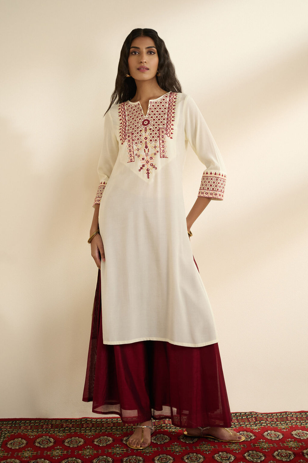 Woman wearing a white kurta with red embroidery and maroon pants on a plain background