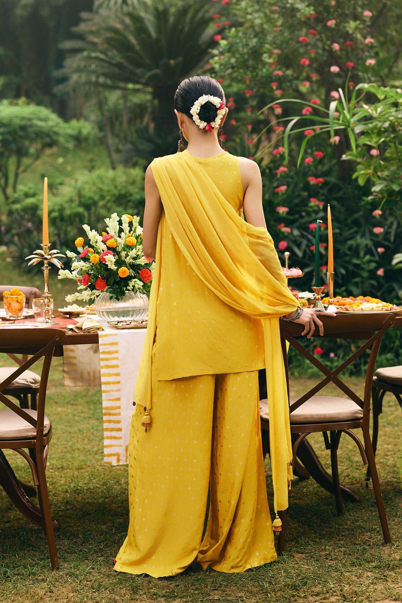 Woman in a yellow saree standing in a garden with tables set for a meal.