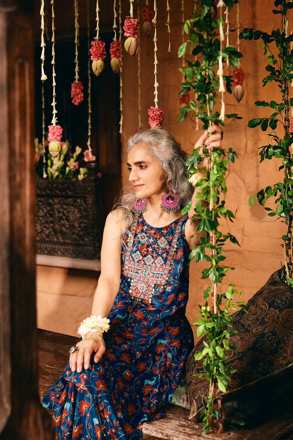 Woman in a patterned dress sitting in a decorated indoor setting with hanging plants and flowers.