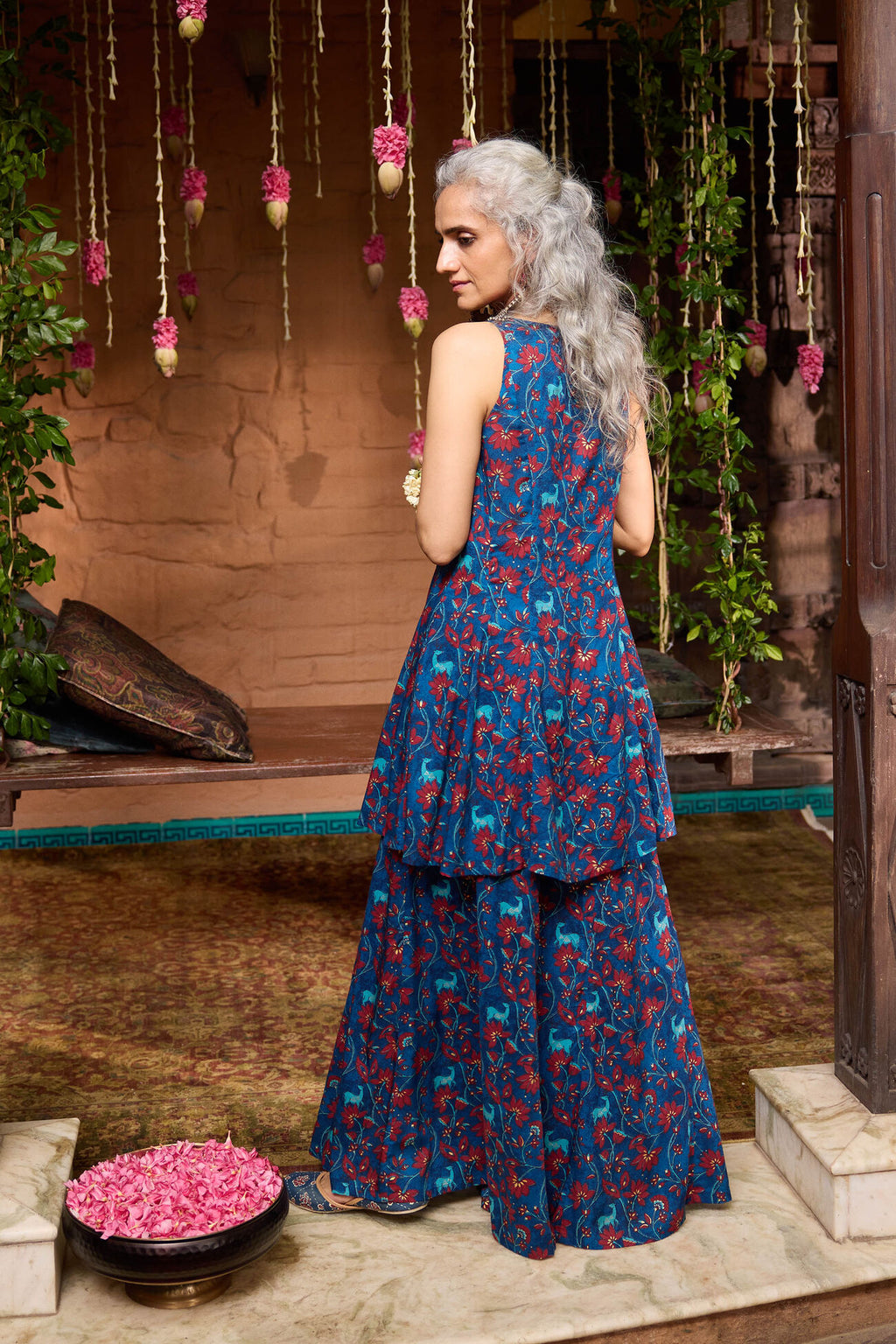 Woman in a blue floral dress standing in a decorative setting with hanging flowers and plants.