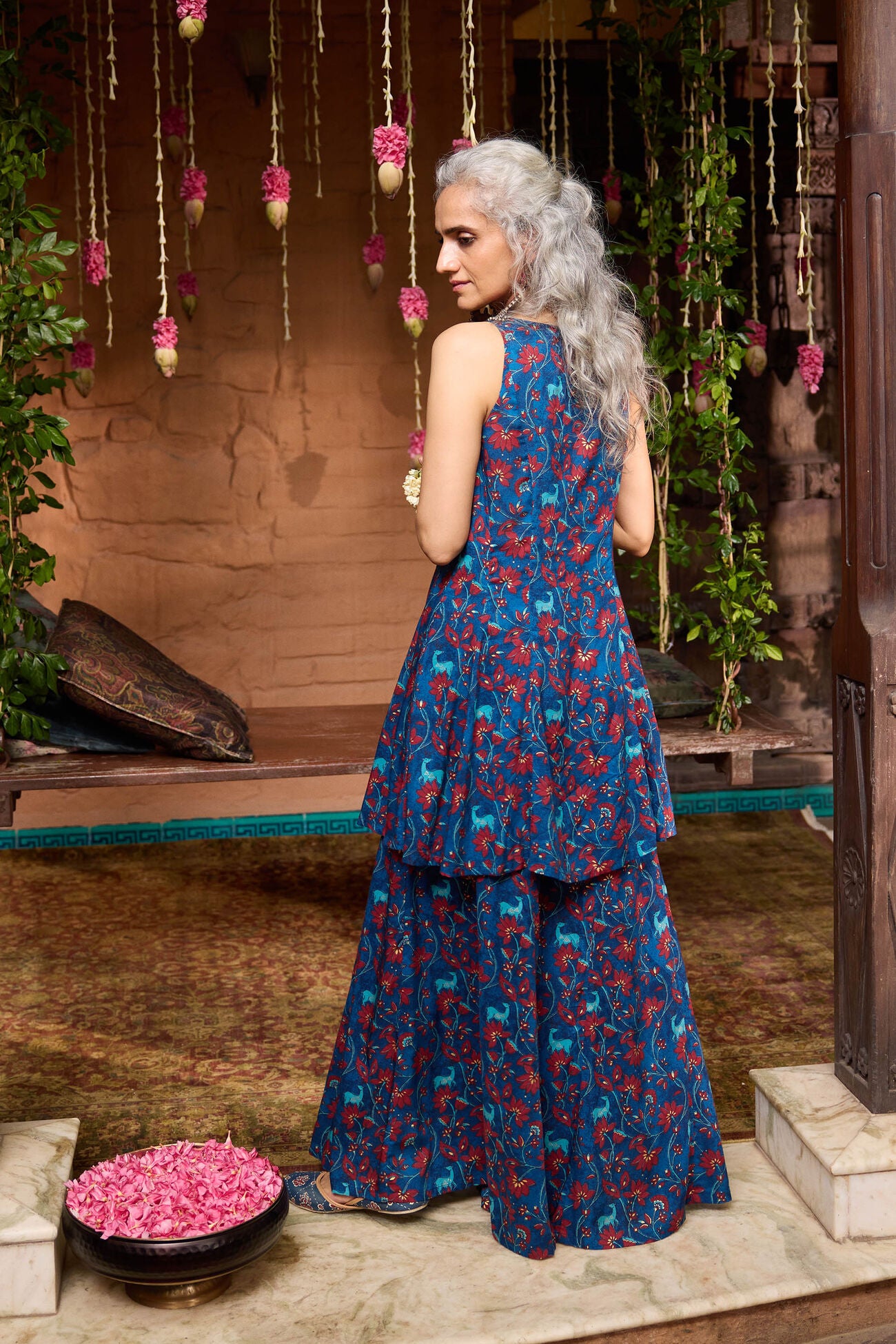 Woman in a blue floral dress standing in a decorative setting with hanging flowers and plants.