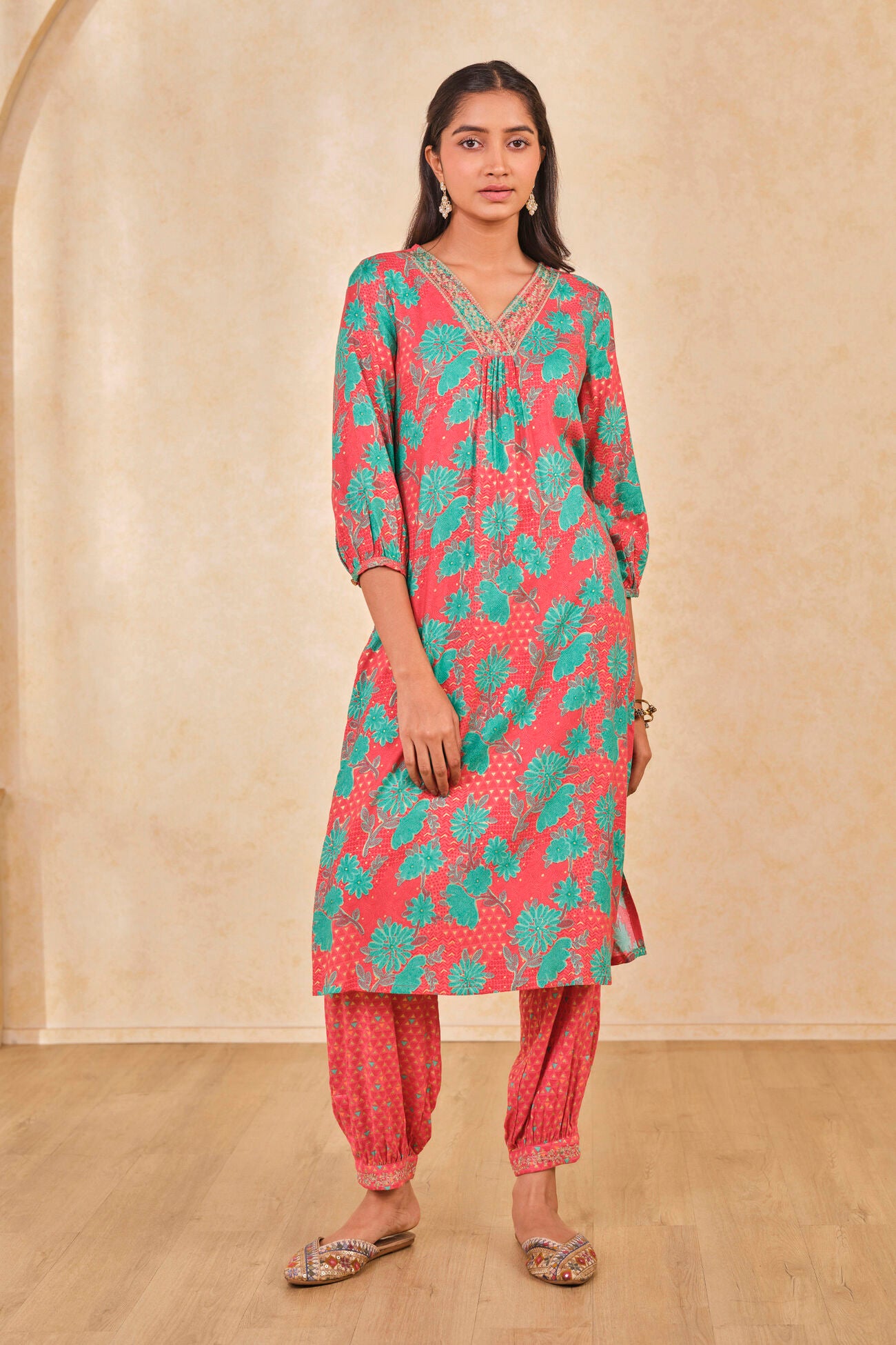 Woman wearing a pink and green patterned kurta with red pants against a beige wall.