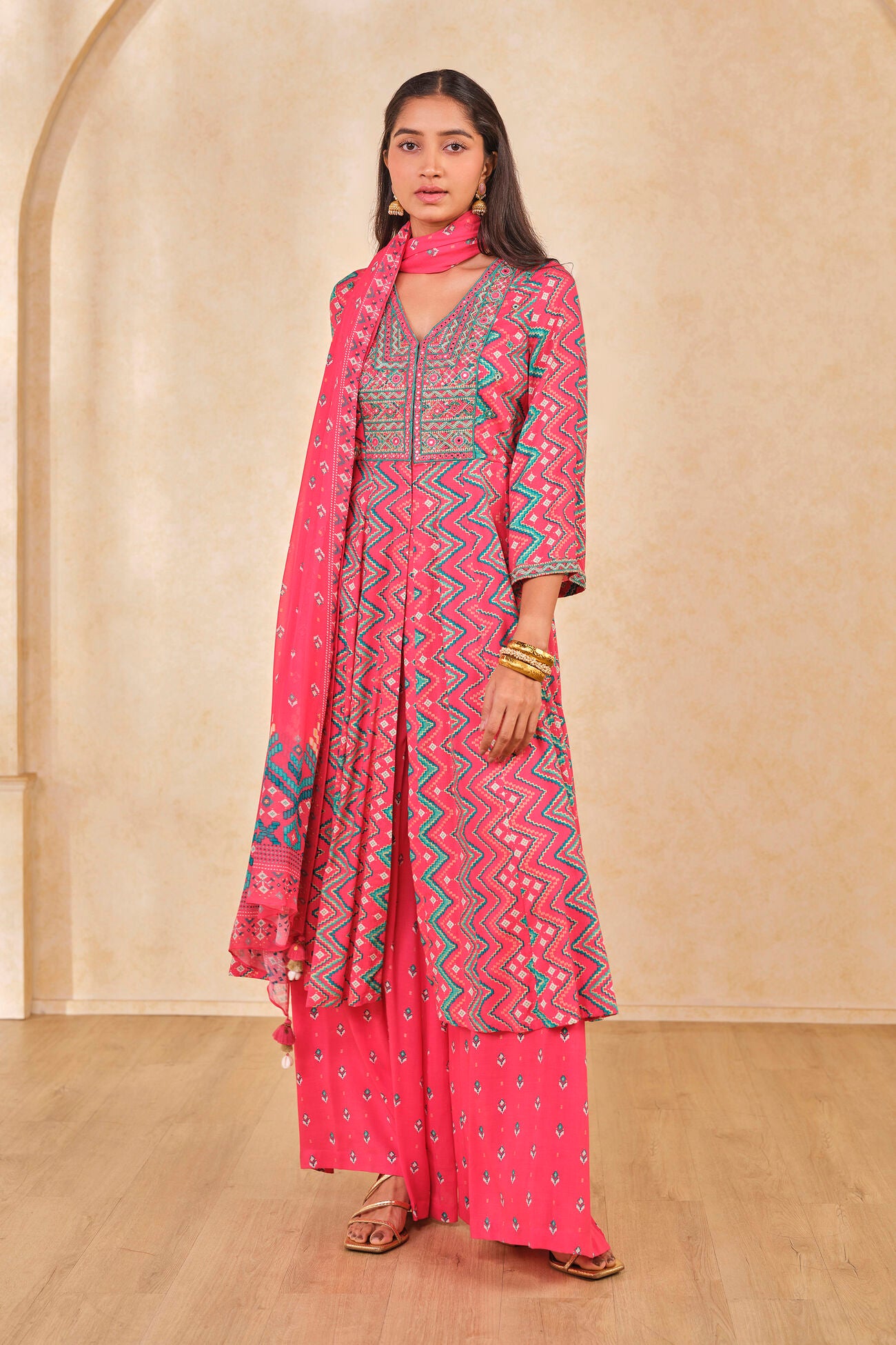 Woman wearing a pink traditional outfit with a patterned dupatta against a beige wall.