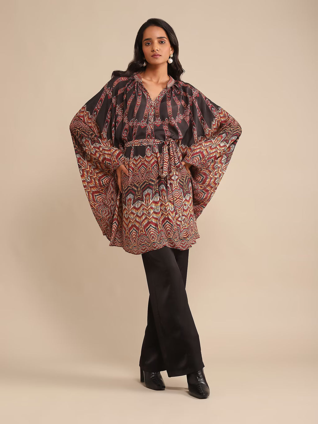Woman in a printed kaftan-style tunic with flared sleeves, paired with black pants and heels, posing against a beige background.