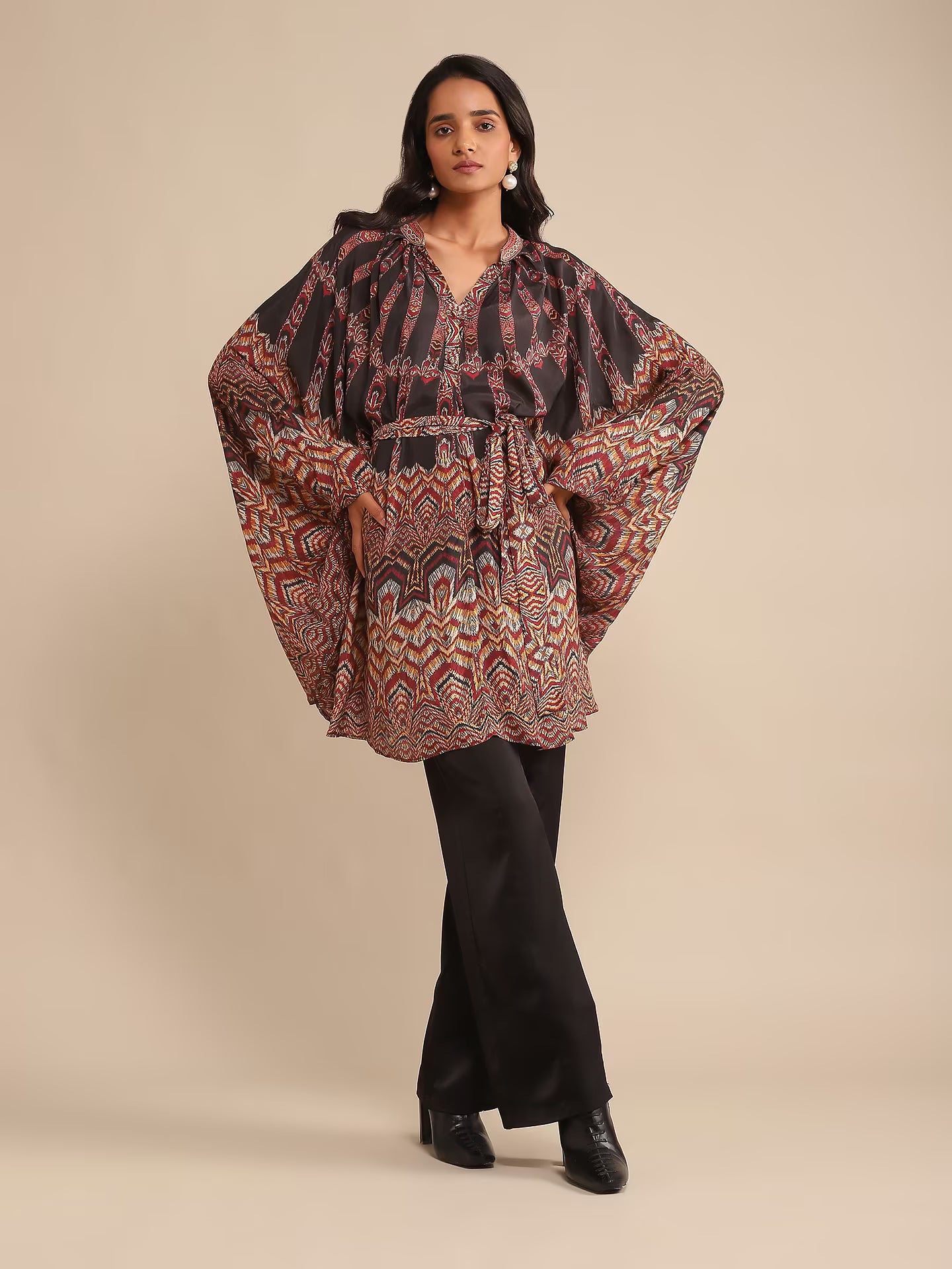 Woman in a printed kaftan-style tunic with flared sleeves, paired with black pants and heels, posing against a beige background.