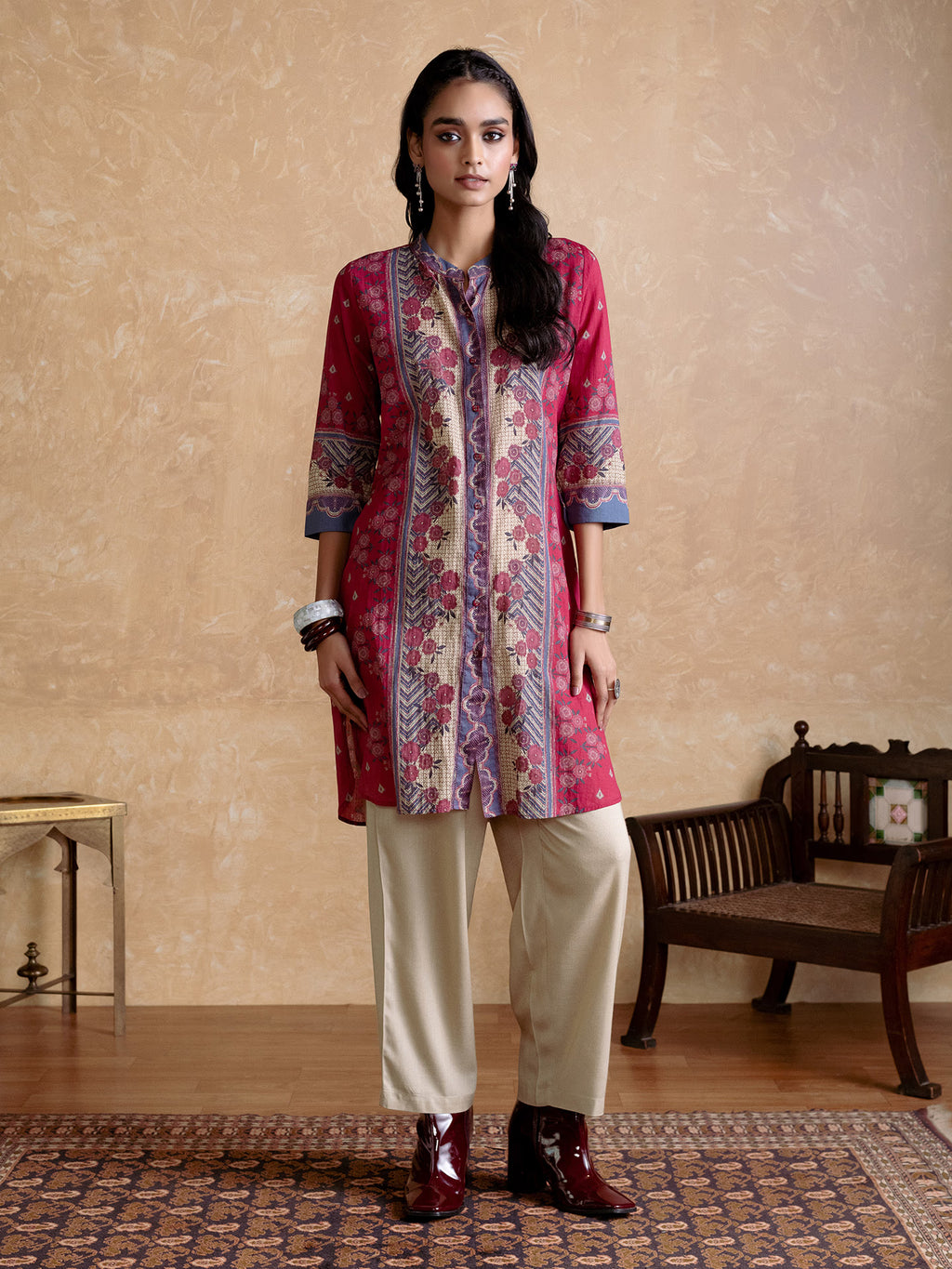 Woman wearing a patterned red and blue kurta with white pants in a room with a wooden bench and table.