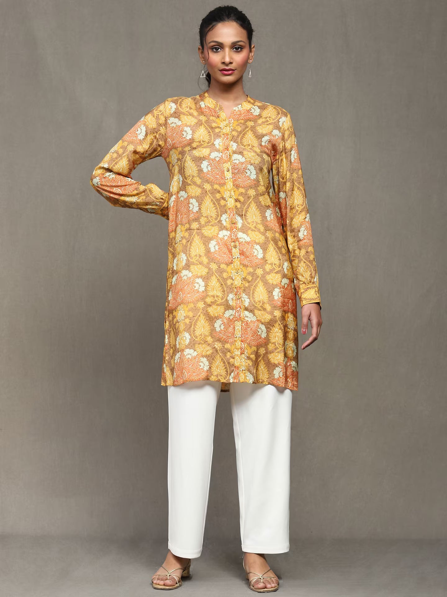 Woman wearing a mustard floral printed kurta with a tan belt, paired with white straight pants and heels, standing against a plain grey background.