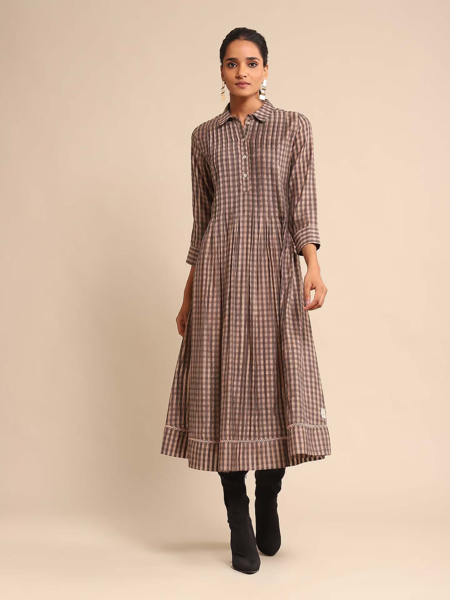 Woman in a brown and beige checked A-line kurta dress with a shirt collar and three-quarter sleeves, paired with black boots, standing against a beige background