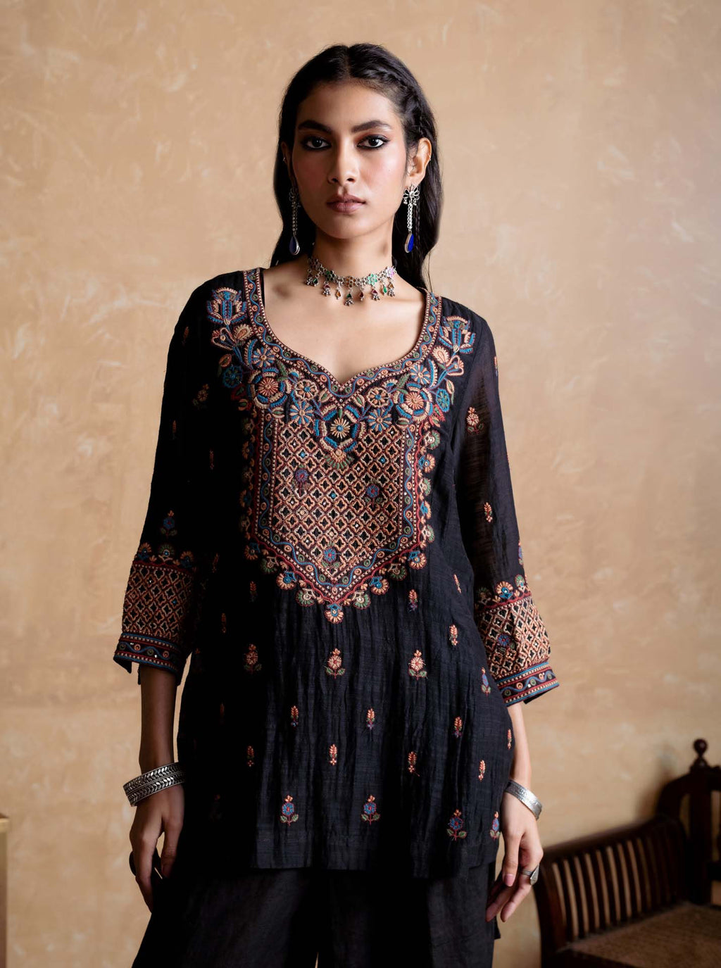 Woman wearing a black embroidered traditional outfit against a beige wall.