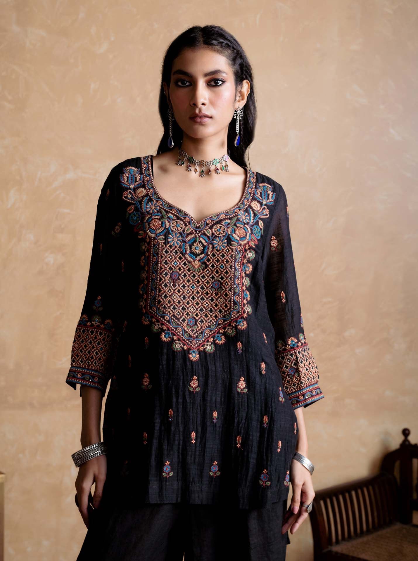 Woman wearing a black embroidered traditional outfit against a beige wall.