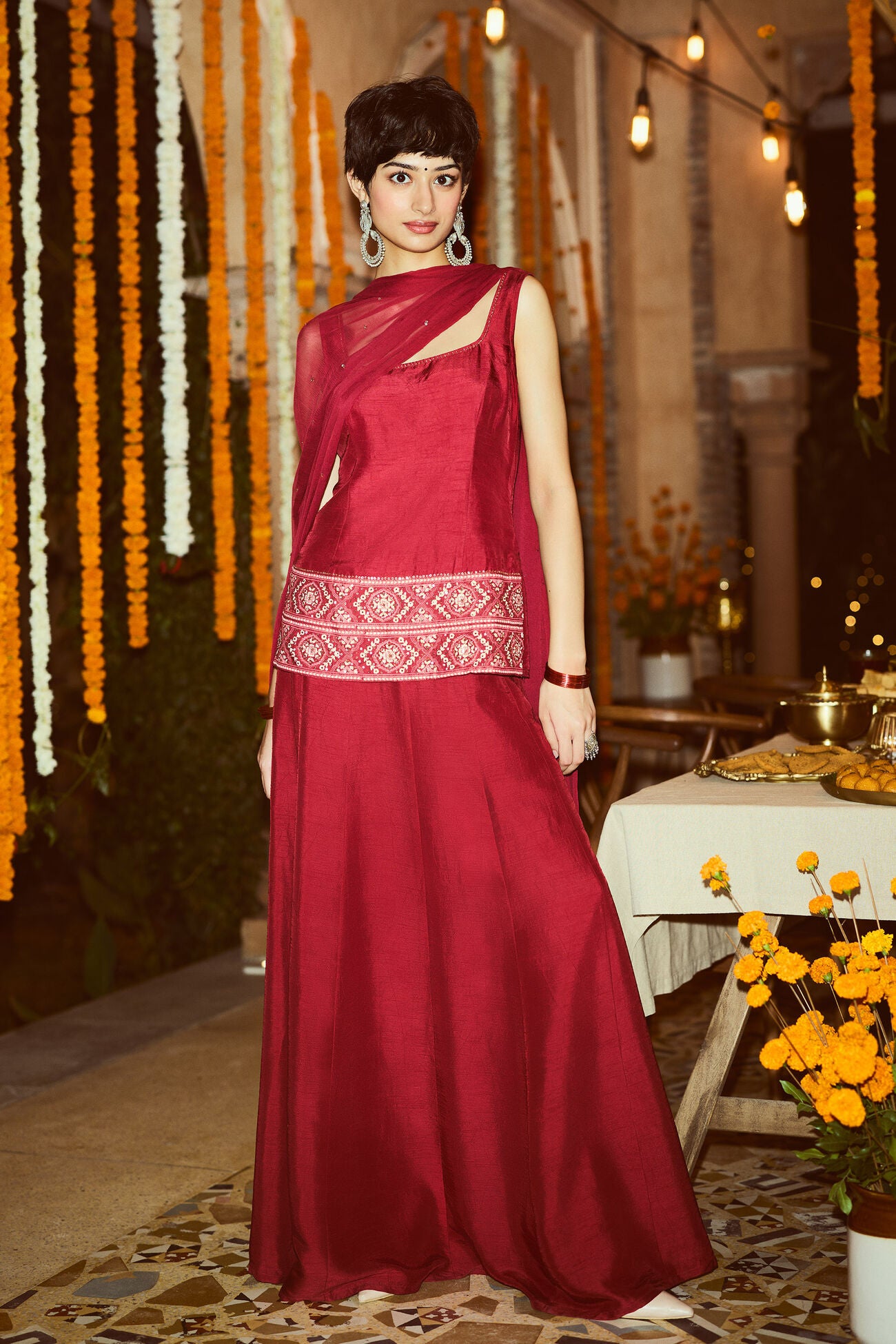 Woman in a red kurta set with a white border standing in a decorated indoor setting.