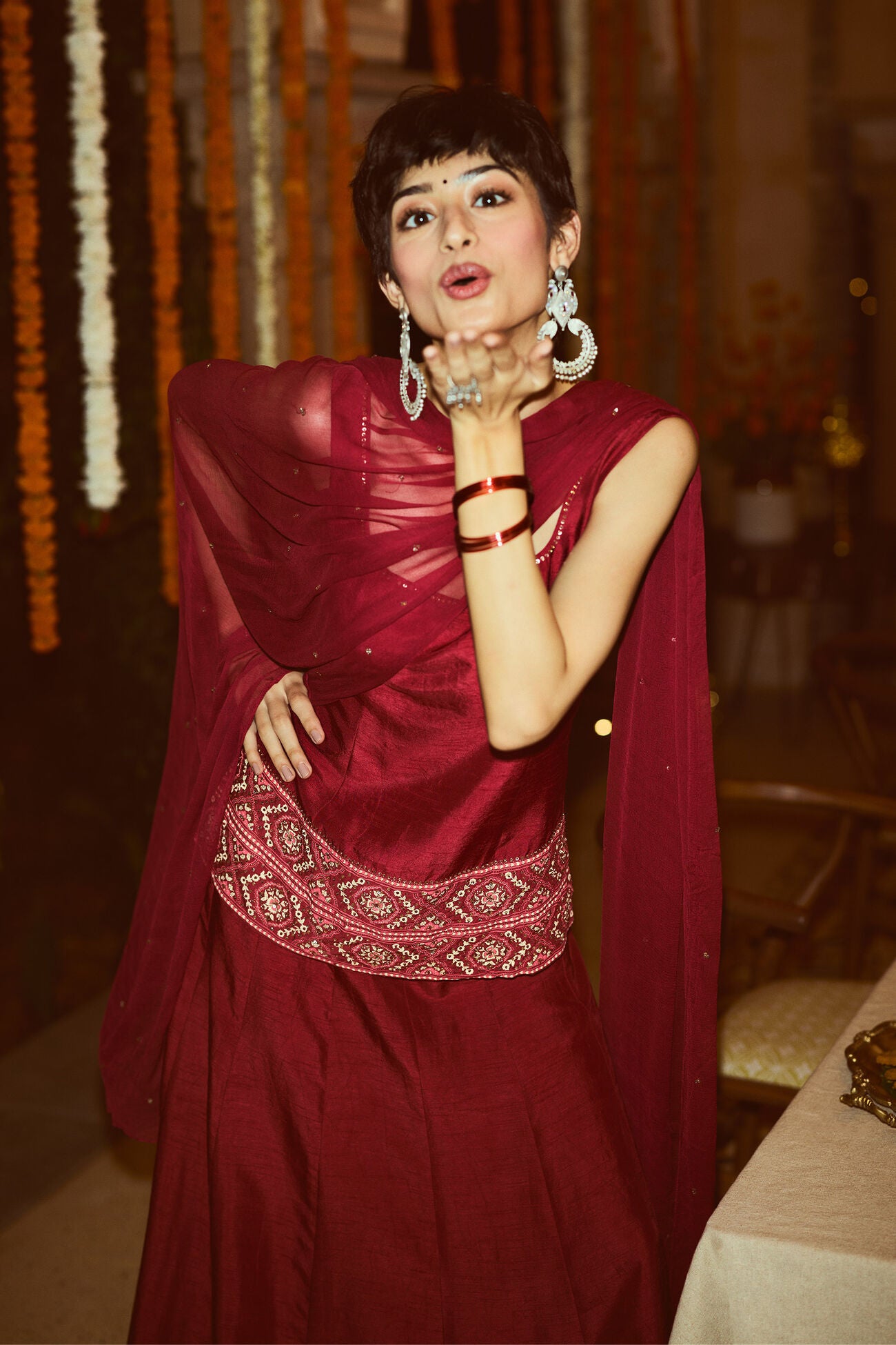 Woman in a red kurta set with a white border standing in a decorated indoor setting.