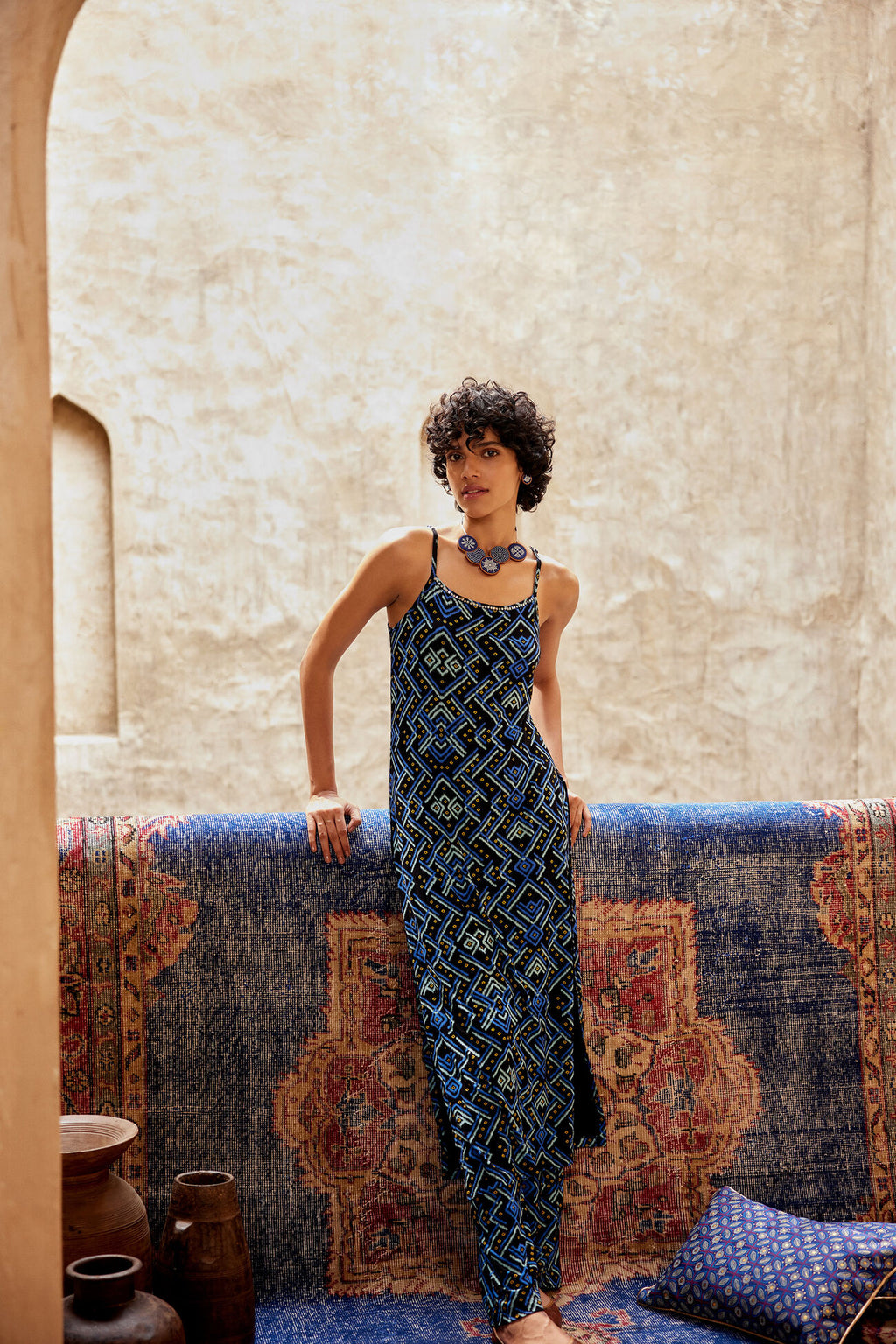 Woman in a blue patterned dress standing in a room with a textured wall and colorful rug.