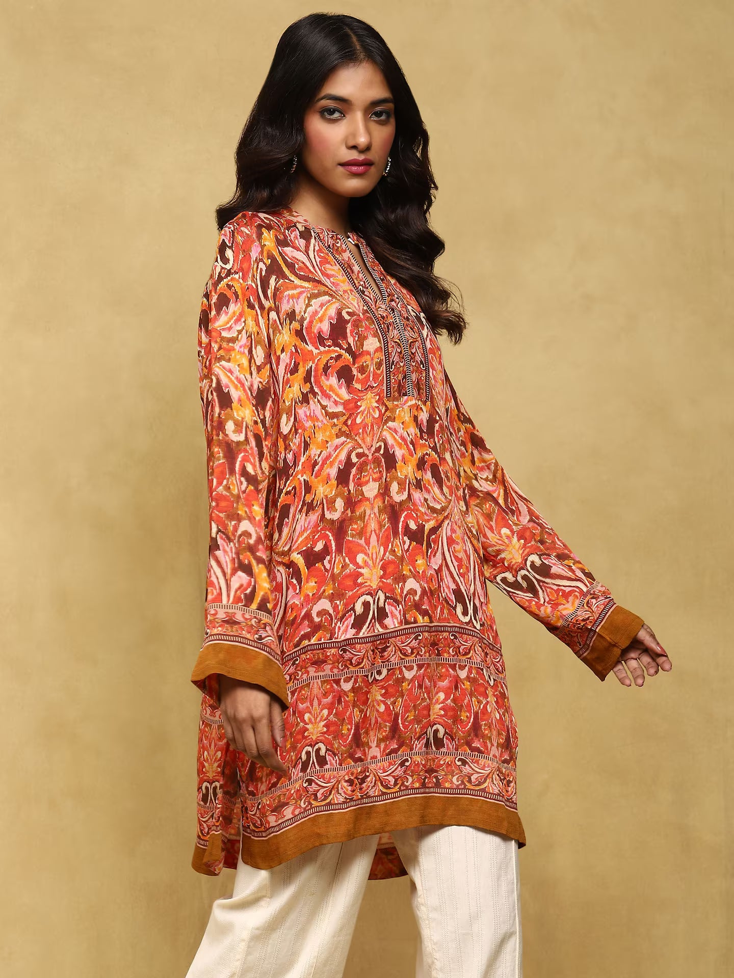 Woman wearing a patterned orange and brown shirt with white pants on a beige background