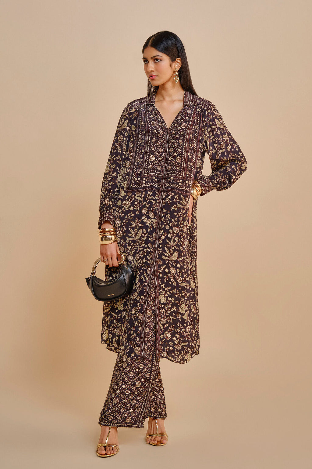 Woman wearing a patterned outfit holding a black handbag against a beige background