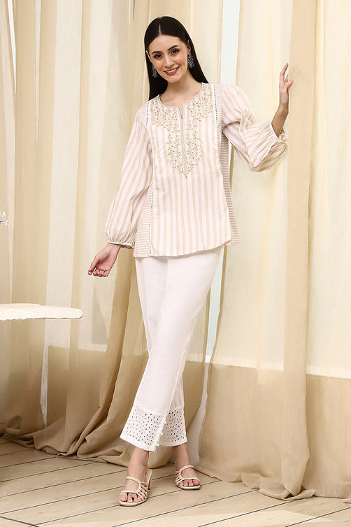 Woman wearing a white and beige outfit with embroidery, standing in front of beige curtains.