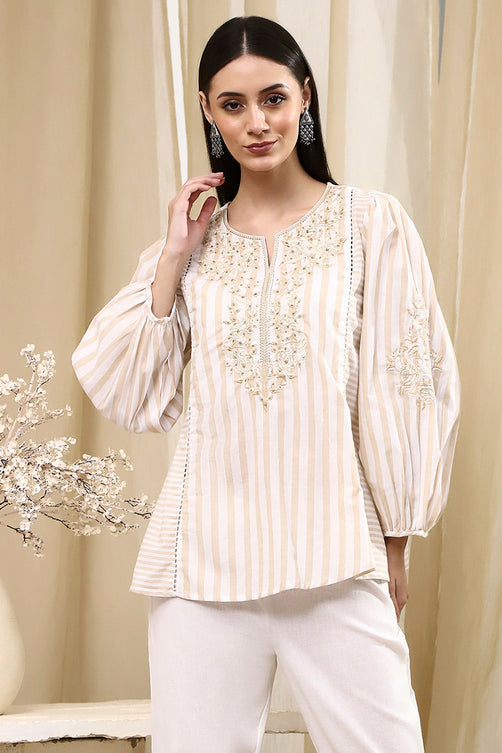 Woman wearing a cream embroidered blouse with white pants against a beige curtain background