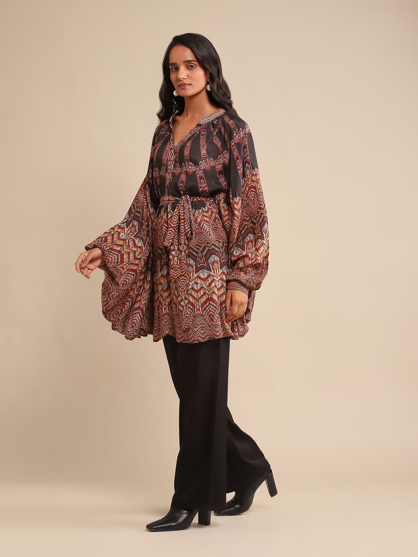 Woman in a printed kaftan-style tunic with flared sleeves, paired with black pants and heels, posing against a beige background.
