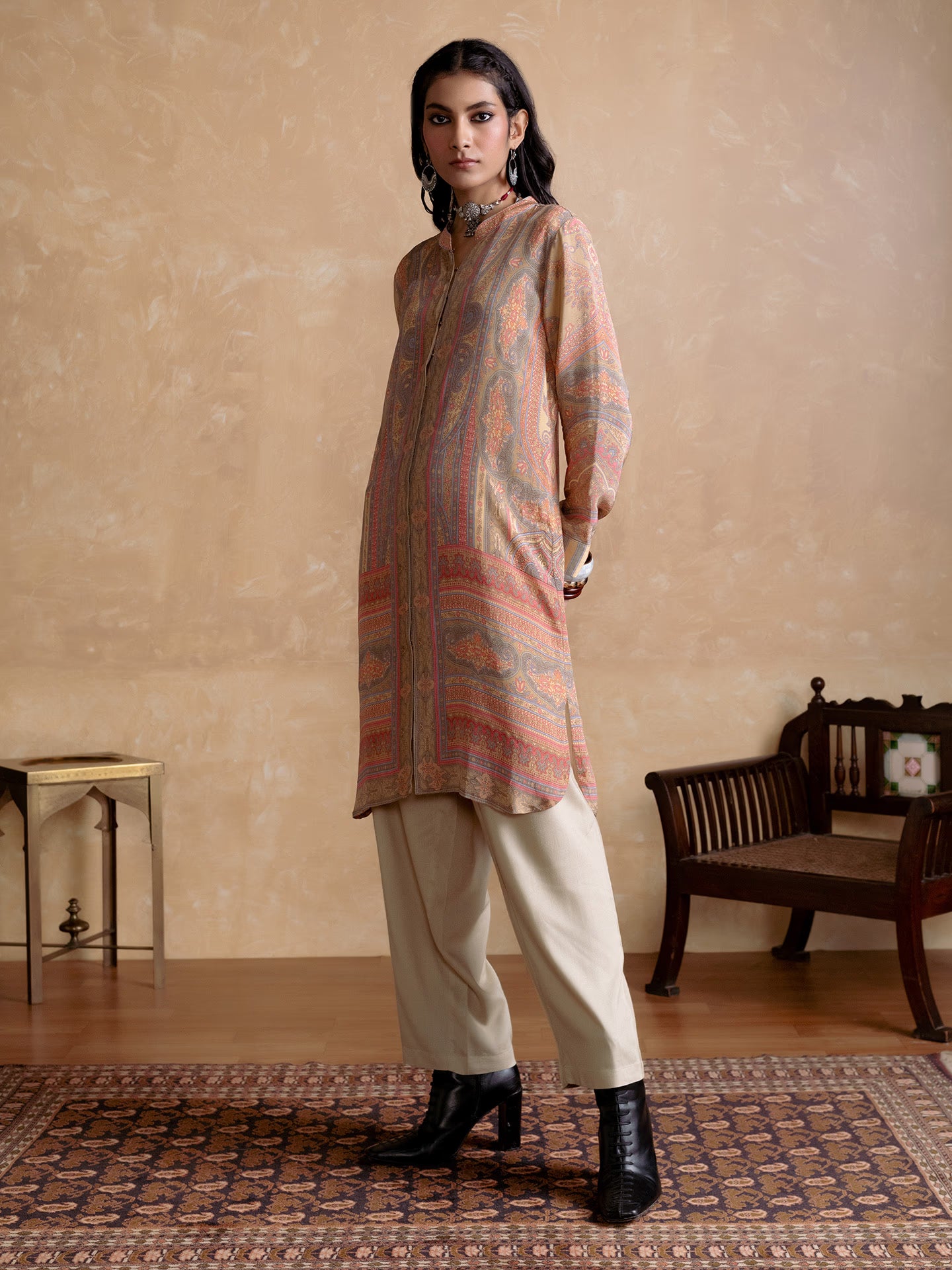 Woman wearing a patterned kurta with beige pants in a room with a wooden bench and small table.