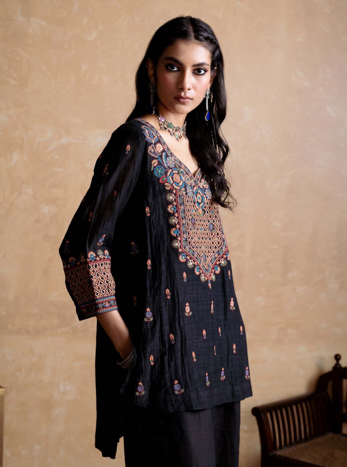 Woman wearing a black embroidered traditional outfit against a beige wall.