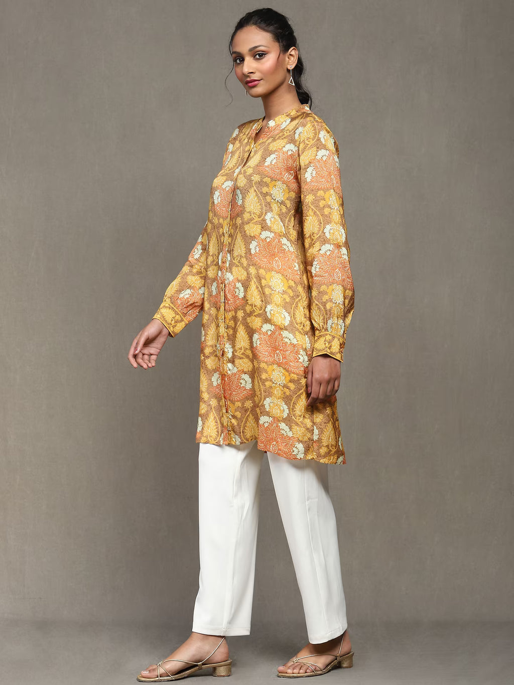 Woman wearing a mustard floral printed kurta with a tan belt, paired with white straight pants and heels, standing against a plain grey background.