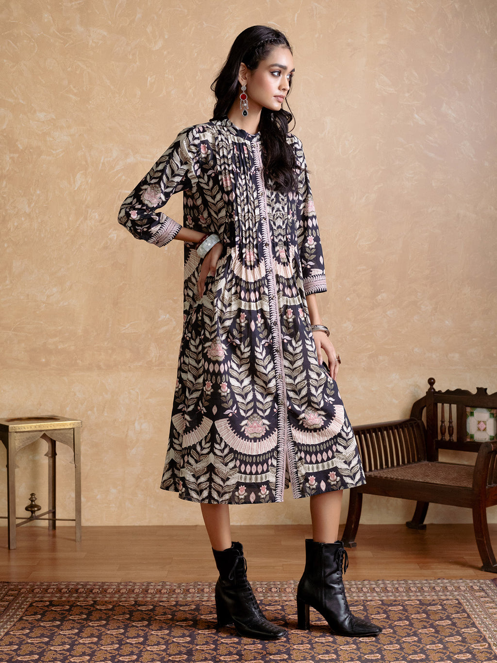 Woman wearing a patterned dress with black boots in a room with a wooden floor and beige walls.