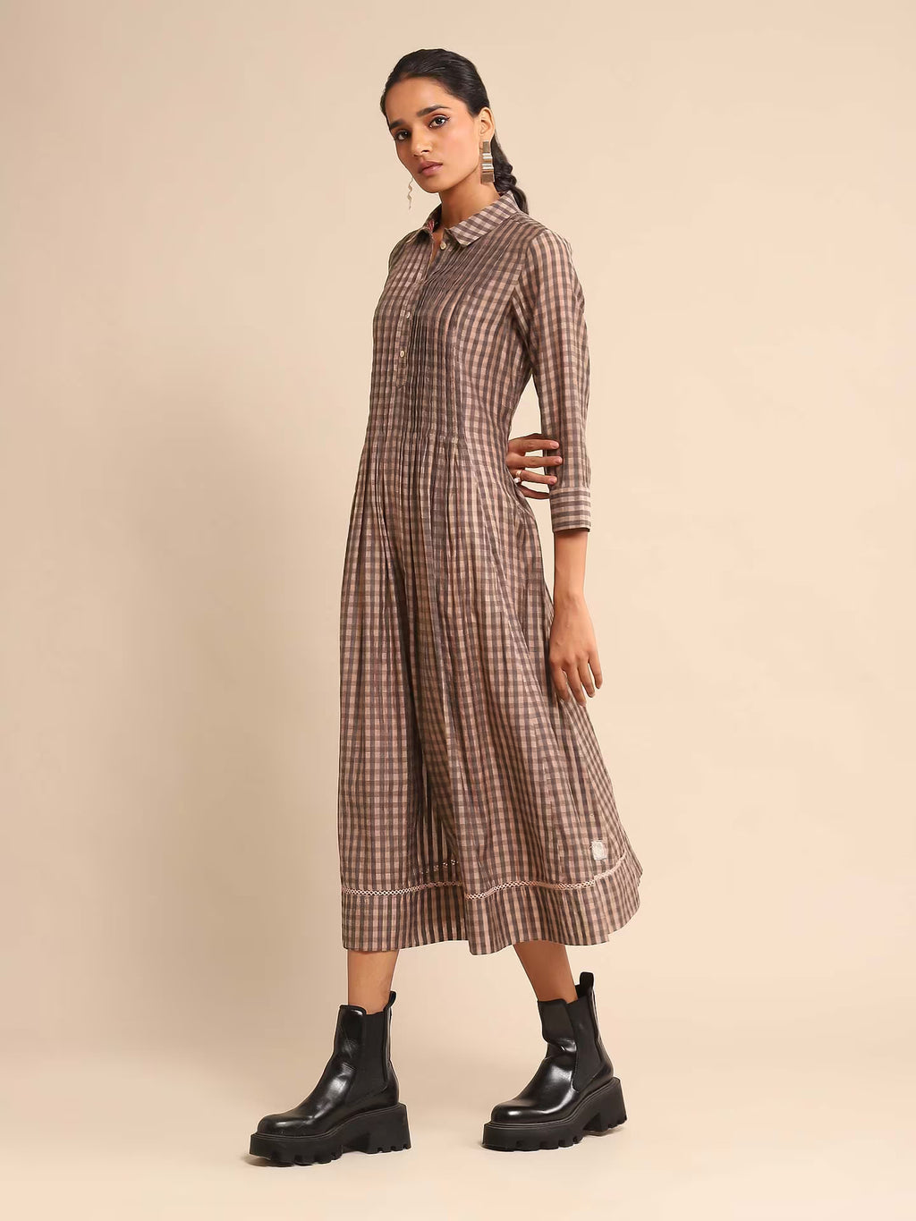 Woman in a brown and beige checked A-line kurta dress with a shirt collar and three-quarter sleeves, paired with black boots, standing against a beige background