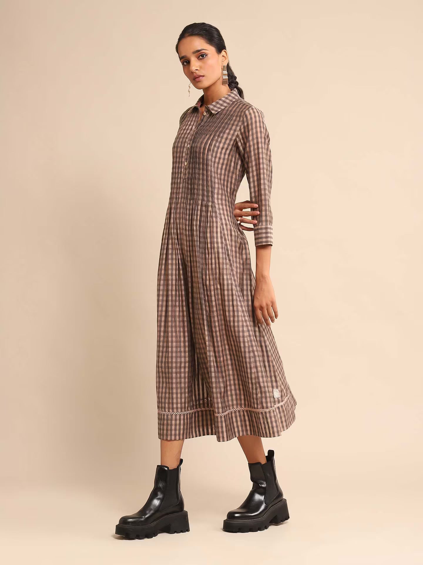 Woman in a brown and beige checked A-line kurta dress with a shirt collar and three-quarter sleeves, paired with black boots, standing against a beige background