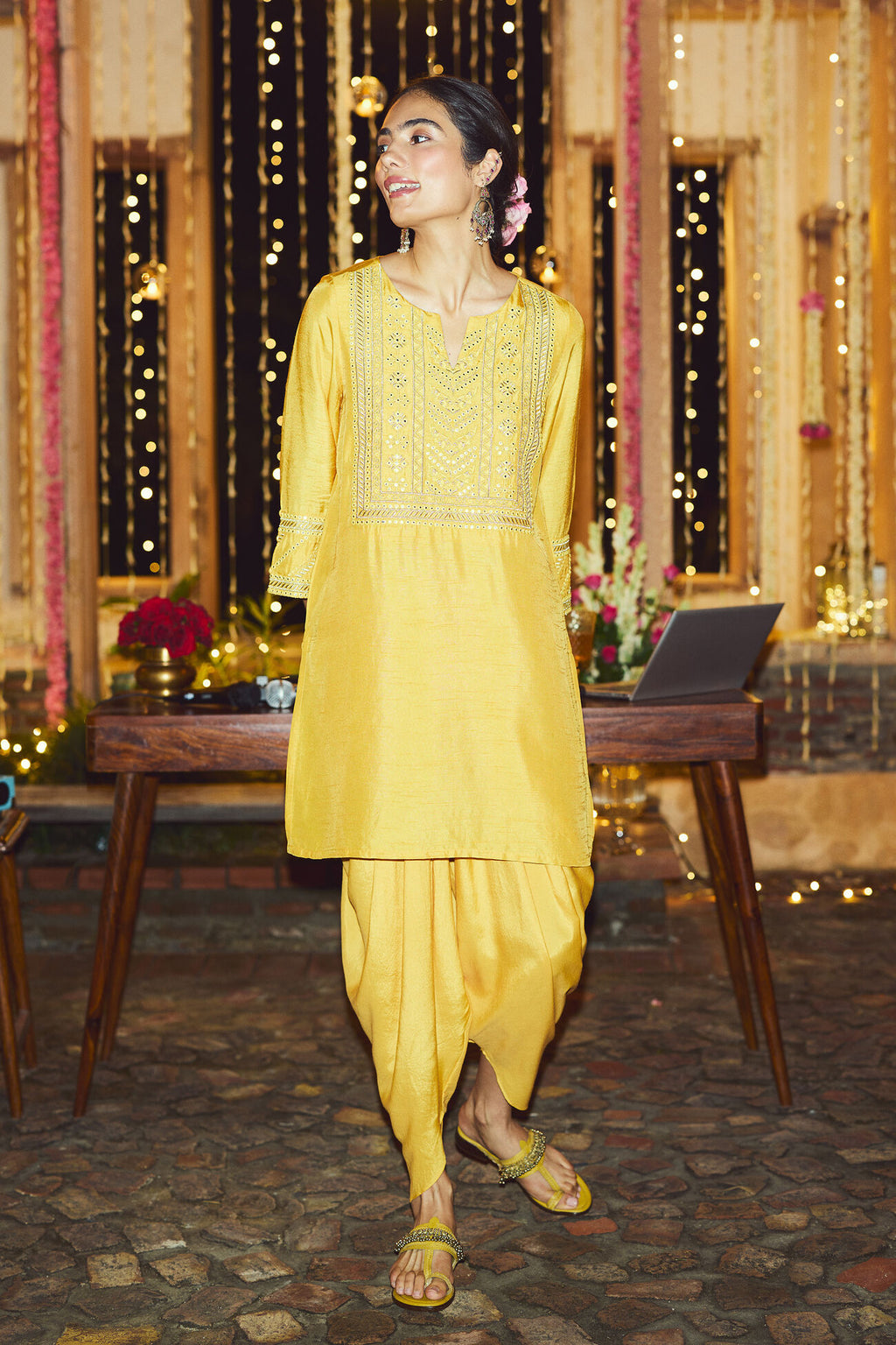 Woman in a yellow traditional outfit standing in a decorated room with lights and flowers.