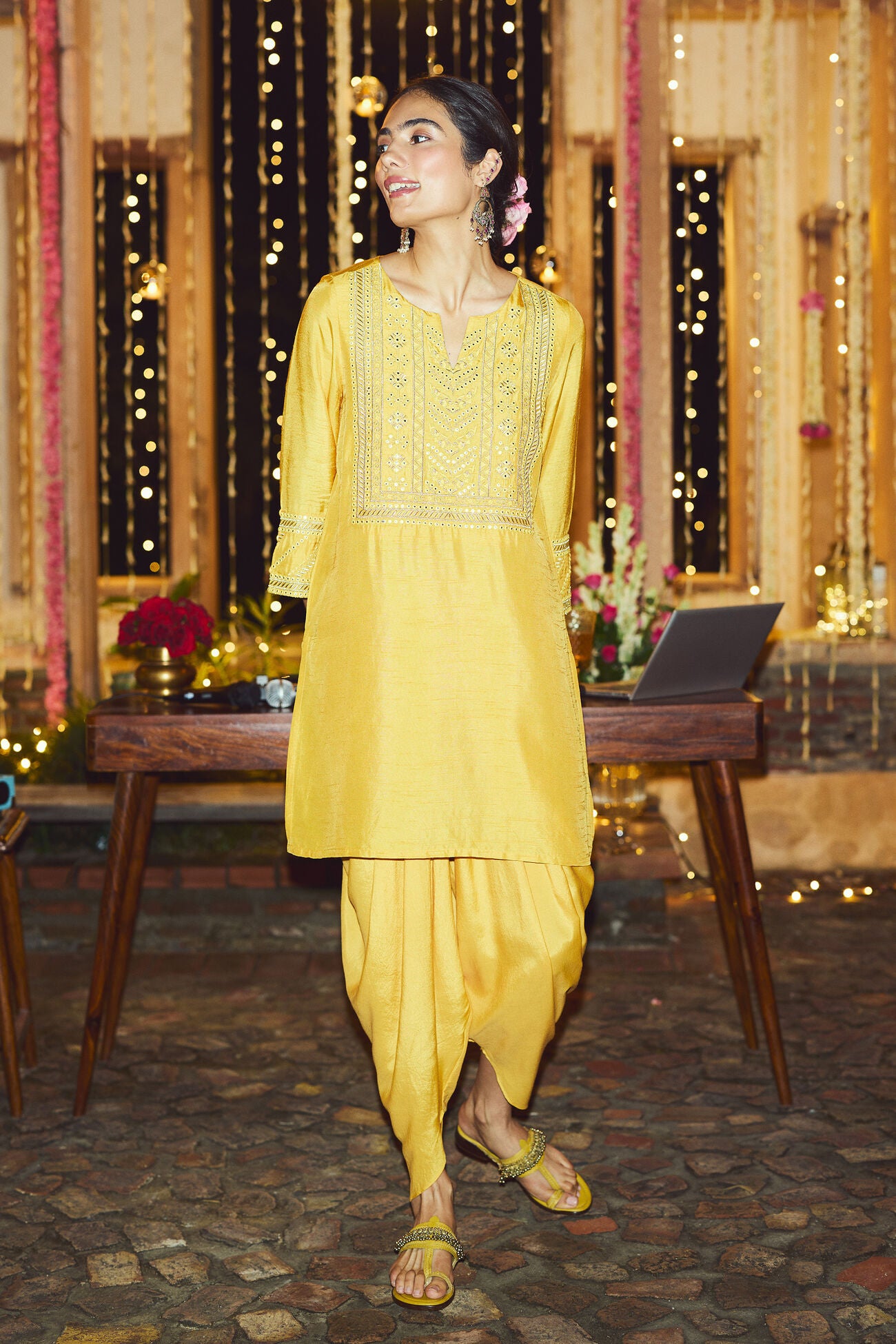 Woman in a yellow traditional outfit standing in a decorated room with lights and flowers.