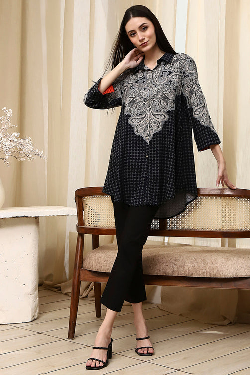 Woman wearing a black and white patterned top and black pants in a room with curtains and a chair.