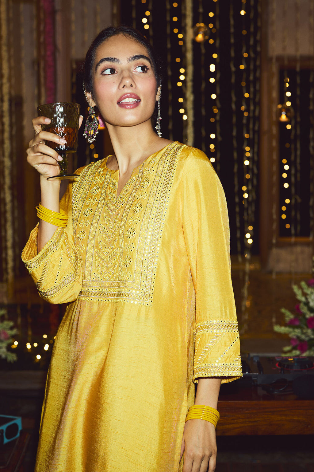 Woman in a yellow traditional outfit holding a glass in a decorated indoor setting