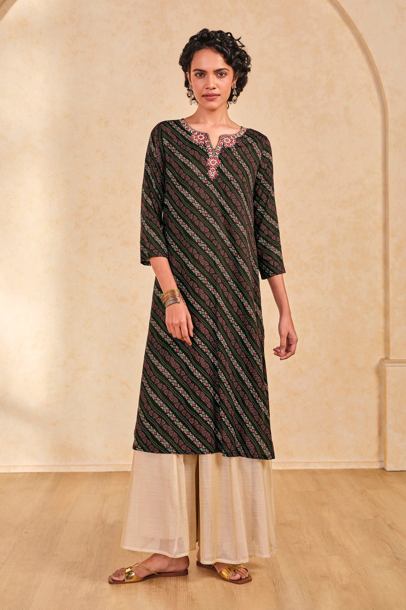Woman wearing a patterned kurta with white pants against a beige wall.