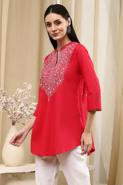 Woman wearing a red embroidered top with white pants in a neutral setting