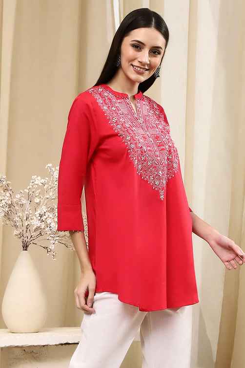 Woman wearing a red embroidered top with white pants in a neutral setting