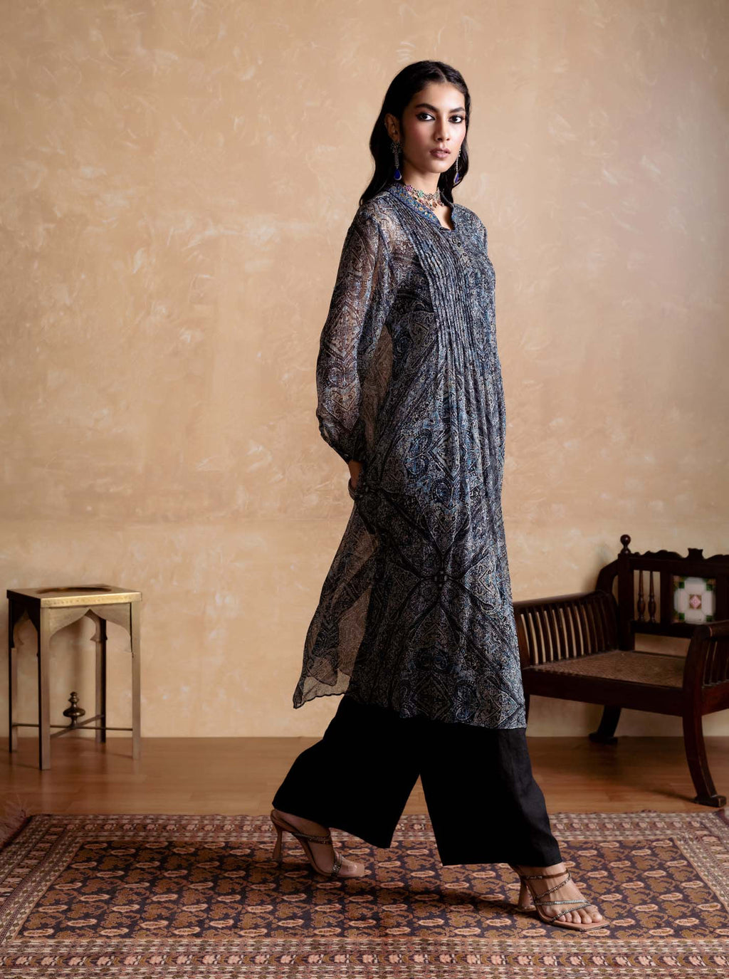Woman wearing a blue and black traditional outfit in a room with a wooden bench and table.