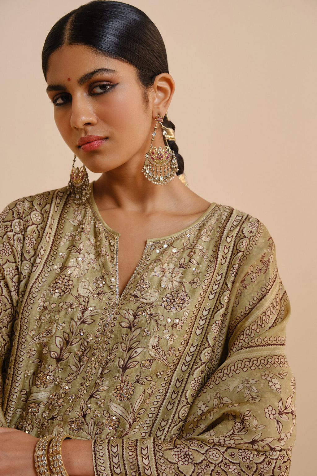 Woman wearing an embroidered traditional outfit with gold jewelry against a beige background