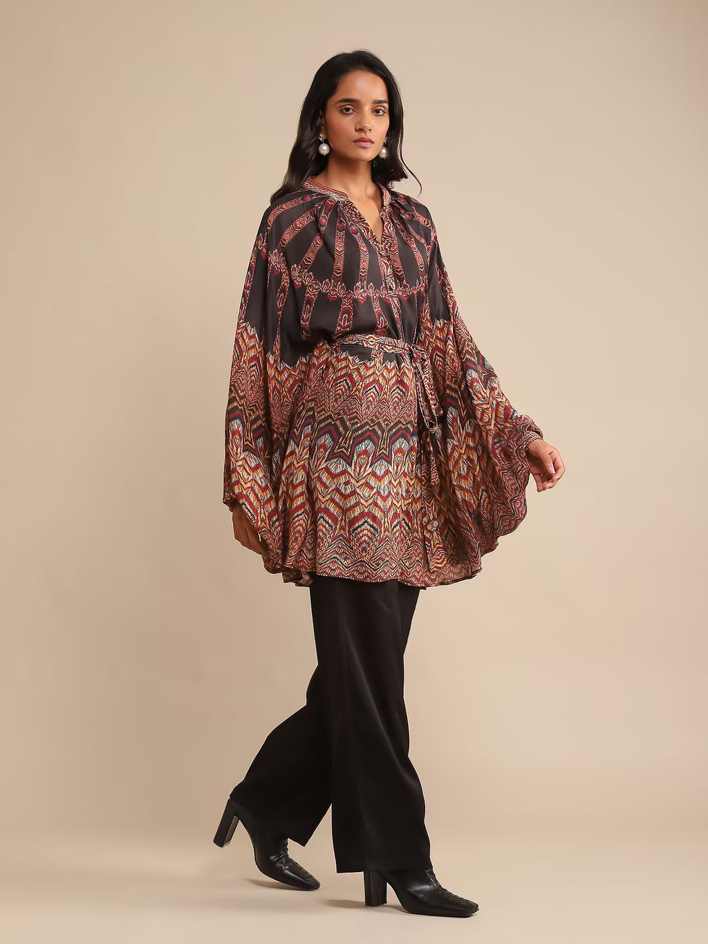Woman in a printed kaftan-style tunic with flared sleeves, paired with black pants and heels, posing against a beige background.