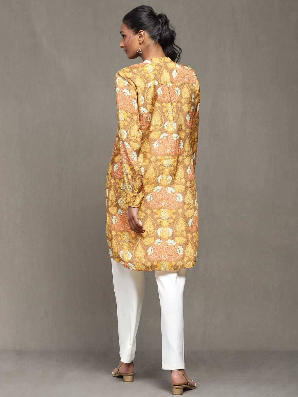 Woman wearing a mustard floral printed kurta with a tan belt, paired with white straight pants and heels, standing against a plain grey background.