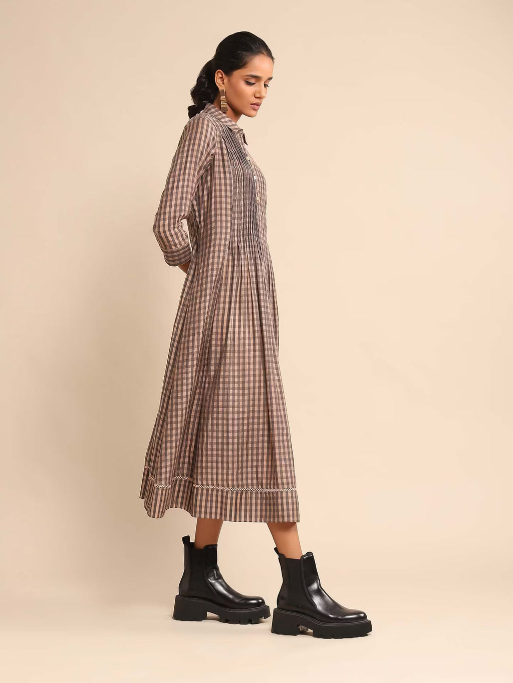 Woman in a brown and beige checked A-line kurta dress with a shirt collar and three-quarter sleeves, paired with black boots, standing against a beige background