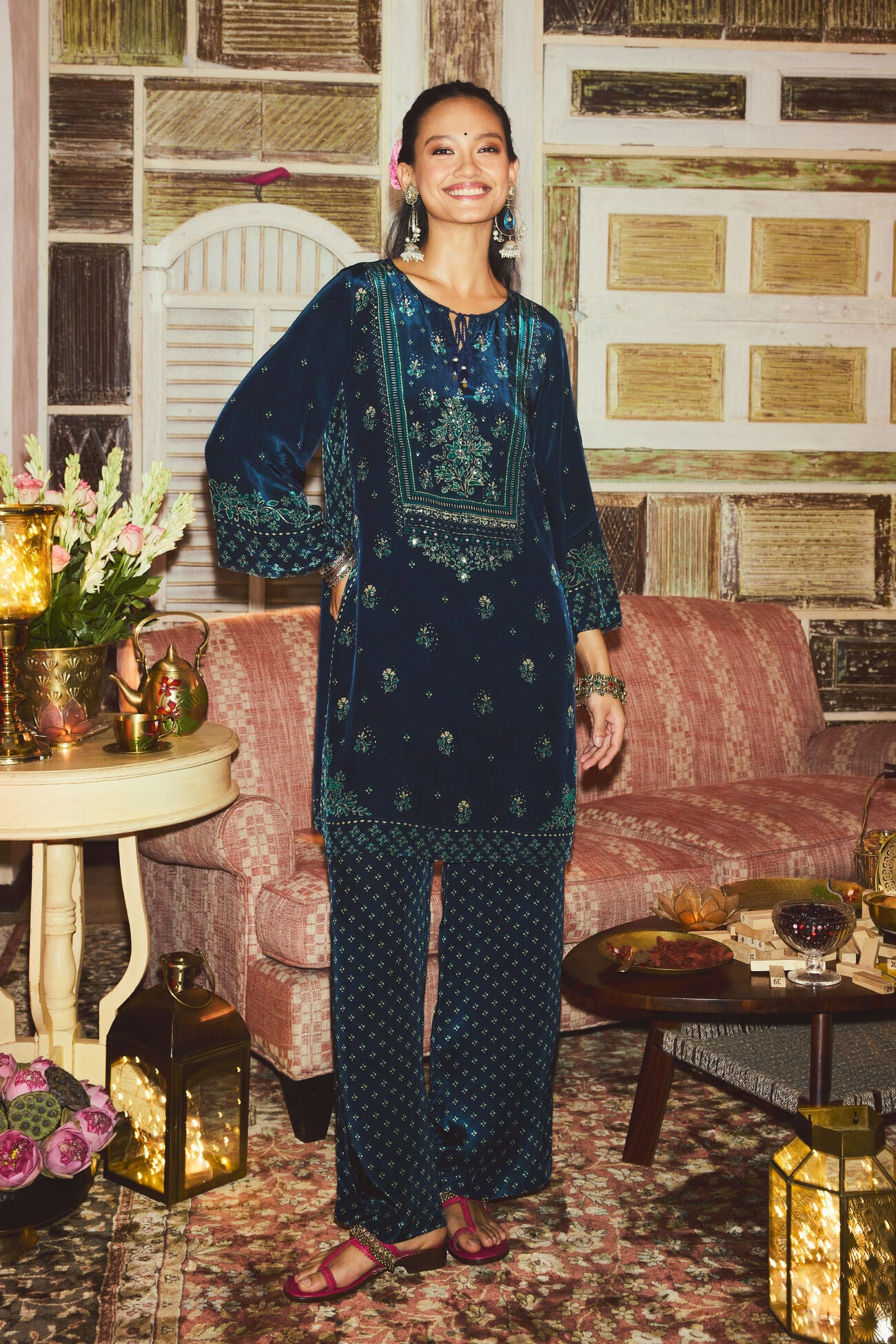 Woman in a dark blue embroidered outfit standing in a decorated room with floral arrangements and lanterns.