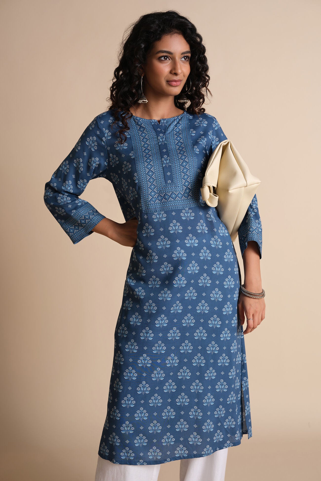 Woman wearing a blue patterned dress holding a beige clutch against a beige background