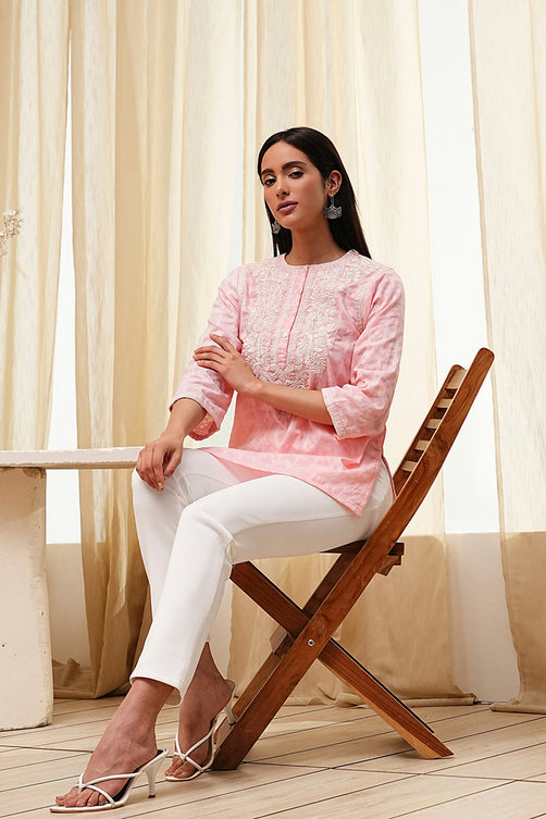 Woman in a pink embroidered top and white pants sitting on a wooden chair with light curtains in the background.