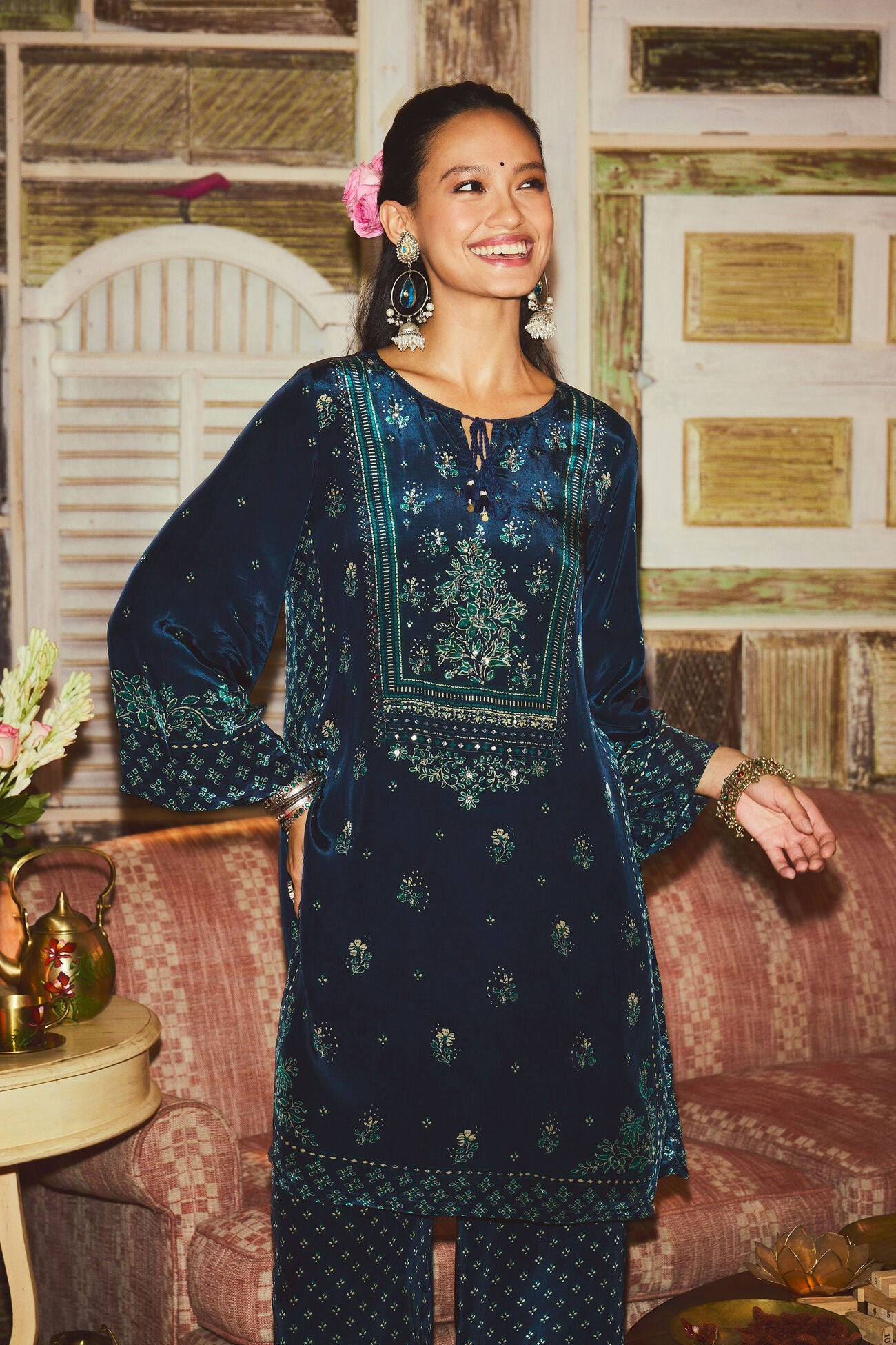 Woman wearing a dark blue embroidered traditional outfit in a decorated room.
