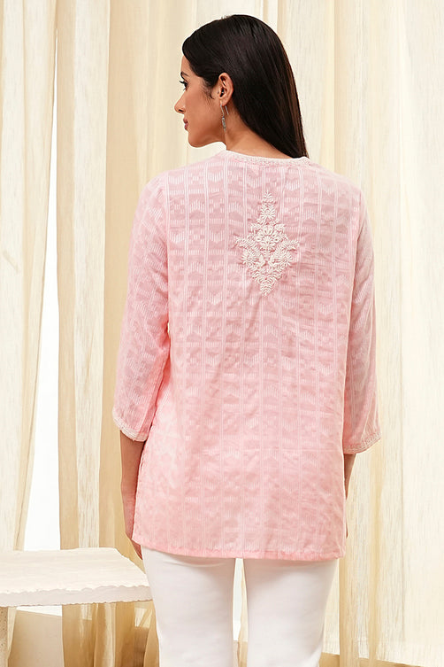 Woman wearing a pink embroidered top with white pants against a beige curtain background