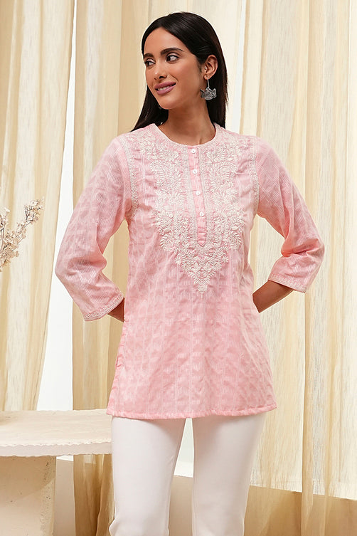 Woman wearing a pink embroidered kurta with white pants against a beige curtain background