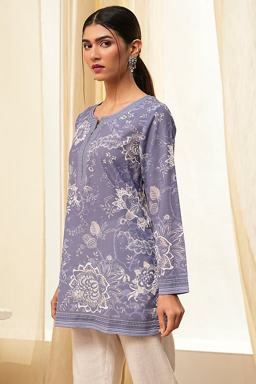 Woman wearing a purple floral patterned shirt with white pants against a light curtain background