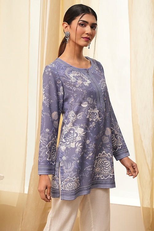 Woman wearing a purple floral embroidered top with beige pants in a bright room.