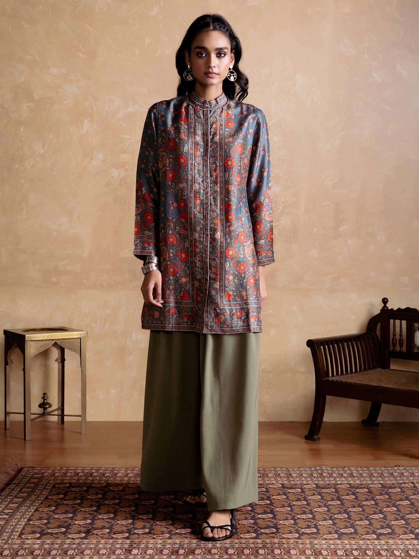 Woman wearing a patterned top and green pants standing in a room with a wooden bench and table.